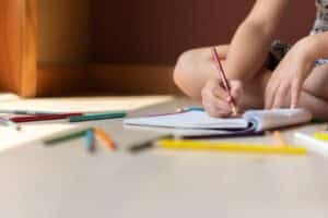 KS2 child sitting on the floor writing a creative story in a notebook surrounded by colouring pencils